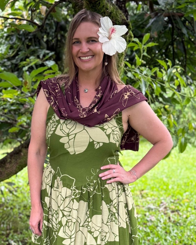 Keolanani (Keola) Kinghorn smiling in a green and cream floral dress with a white hibiscus in her hair, standing in a garden.
